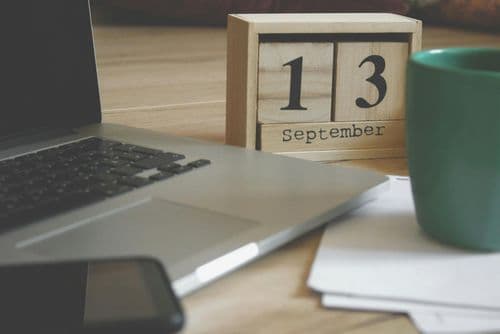 A wooden desk with a laptop, smartphone, green coffee mug, and a wooden block calendar showing September 13.