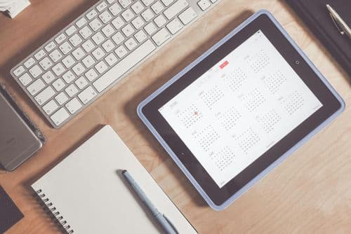 A tablet displaying a digital calendar on a wooden desk beside a keyboard, notebook, and pen — representing calendar-based budgeting and financial planning.